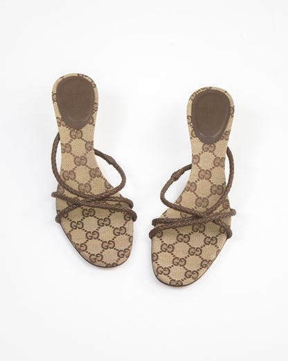 A pair of Gucci flat slide sandals featuring the brand's signature brown and beige GG monogram canvas, with brown leather straps wrapped around the toes, shown against a plain white background.