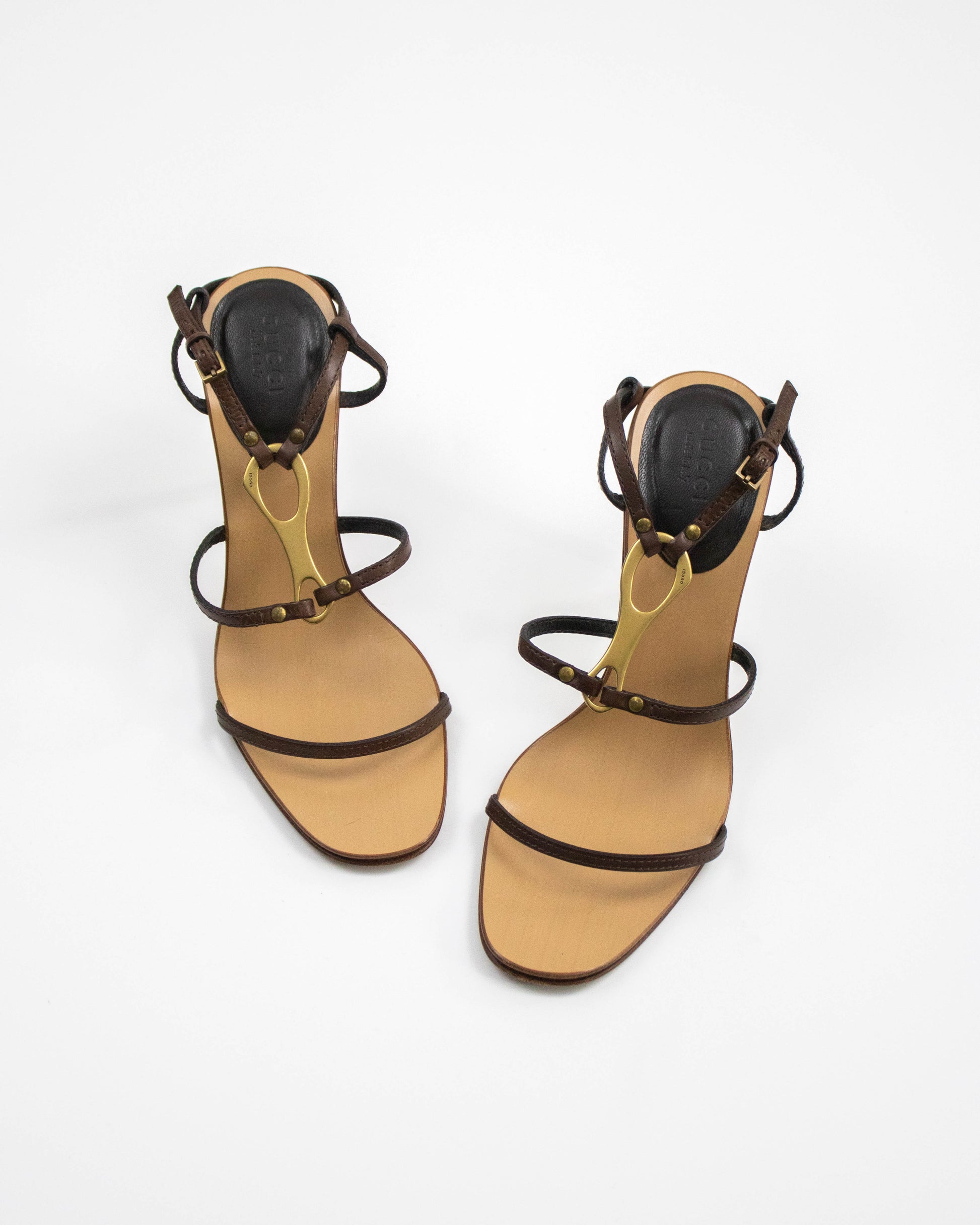 Pair of Gucci high-heeled sandals with brown straps and gold horsebit accents on a white background front view