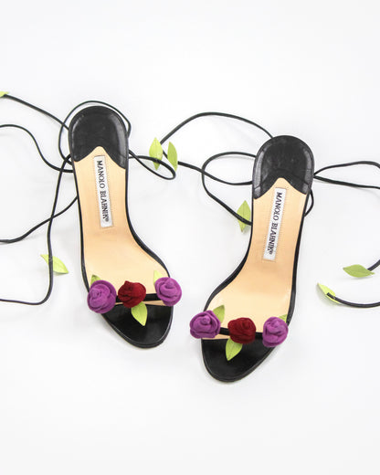 A pair of black high-heeled sandals by Manolo Blahnik, decorated with purple and burgundy fabric roses and green leaves on the toe strap, with black lace-up straps extending across a white background.