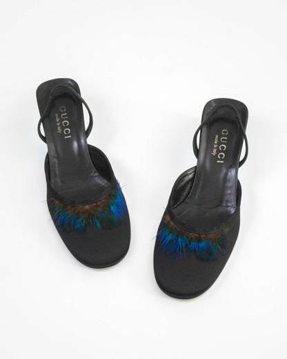 A pair of black Gucci slingback mule sandals with iridescent blue and green feather trim on the toe, shown from a top-down view on a white background.